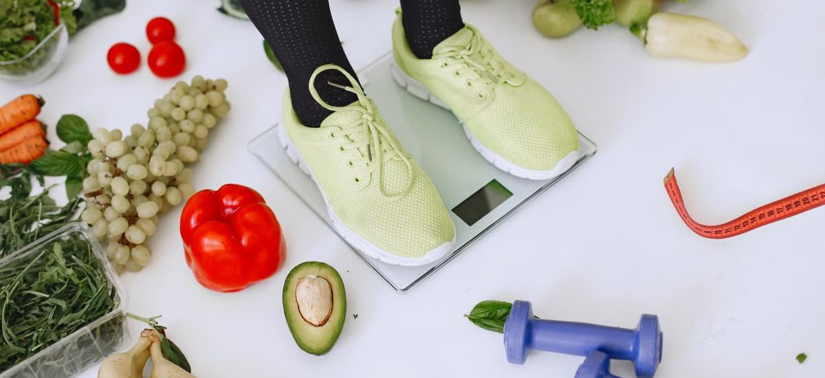 Person standing on scales surrounded by fresh produce and fitness equipment symbolizing a balanced lifestyle.